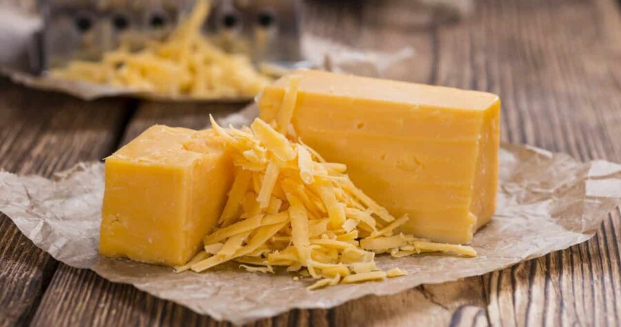 Blocks of yellow cheddar cheese and a small pile of grated cheddar placed on crumpled parchment paper over a rustic wooden table