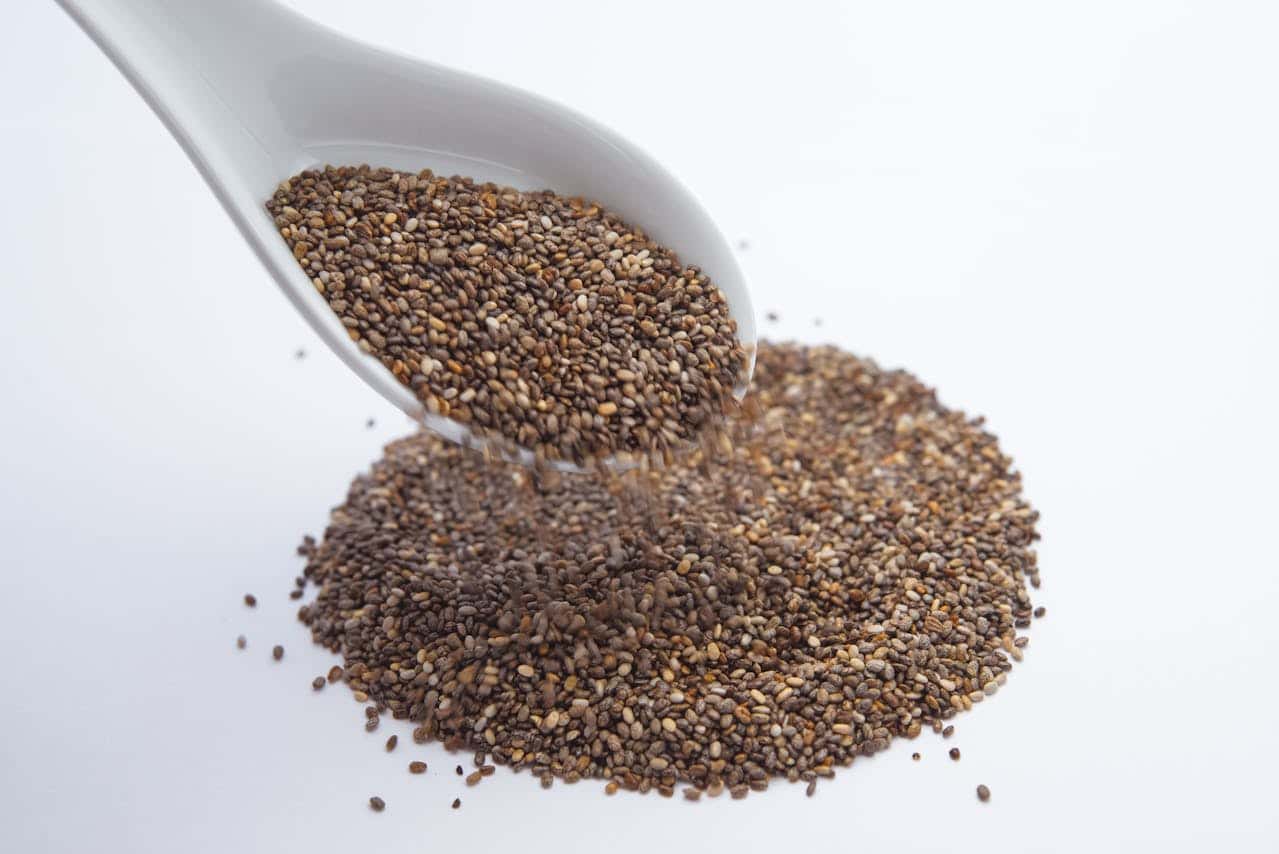 White ceramic spoon pouring chia seeds, small brown and beige seeds falling, pile of chia seeds on a white surface, healthy superfood, close-up shot