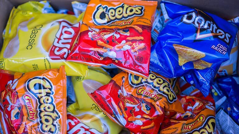 A close-up of several bags of snacks, including Cheetos, Fritos, and other chip varieties, with bright packaging in yellow, orange, and blue, stacked together