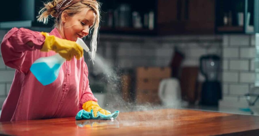 Young woman wearing yellow gloves, spraying an oil-based wood cleaner on a wooden surface, wiping with a cloth, creating a mist effect, cleaning a kitchen countertop, focused expression, cozy home setting
