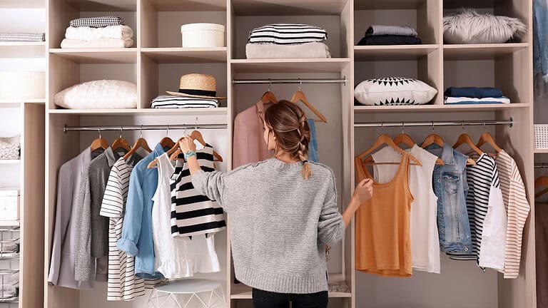 A person browsing through a closet, looking at hanging clothes, organizing outfits, choosing clothes to wear, light-colored and striped shirts visible