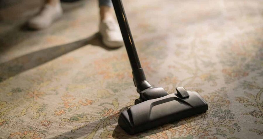 Vacuum cleaner in use on a patterned carpet, person wearing white sneakers partially visible, home cleaning in progress
