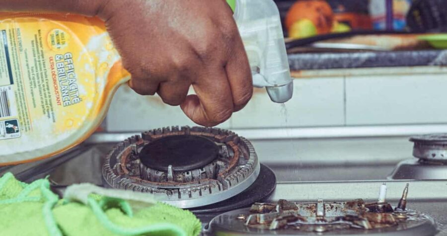 Hand spraying cleaning solution on a greasy gas stove, green microfiber cloth nearby, deep cleaning kitchen appliance, removing grime and stains, household cleaning task, maintaining kitchen hygiene