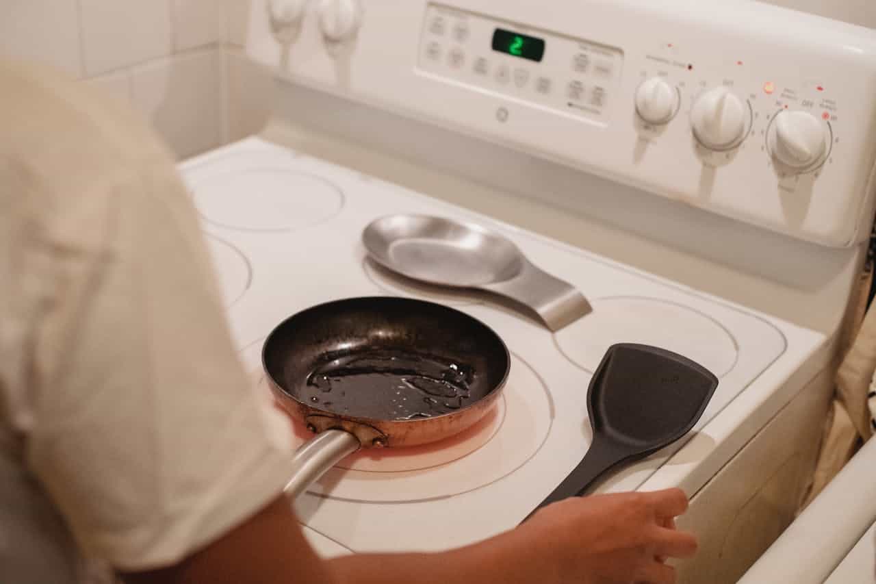 Person cooking on a white electric stove, small frying pan with oil on heated burner, black spatula nearby, digital display showing heat level, spoon rest in background