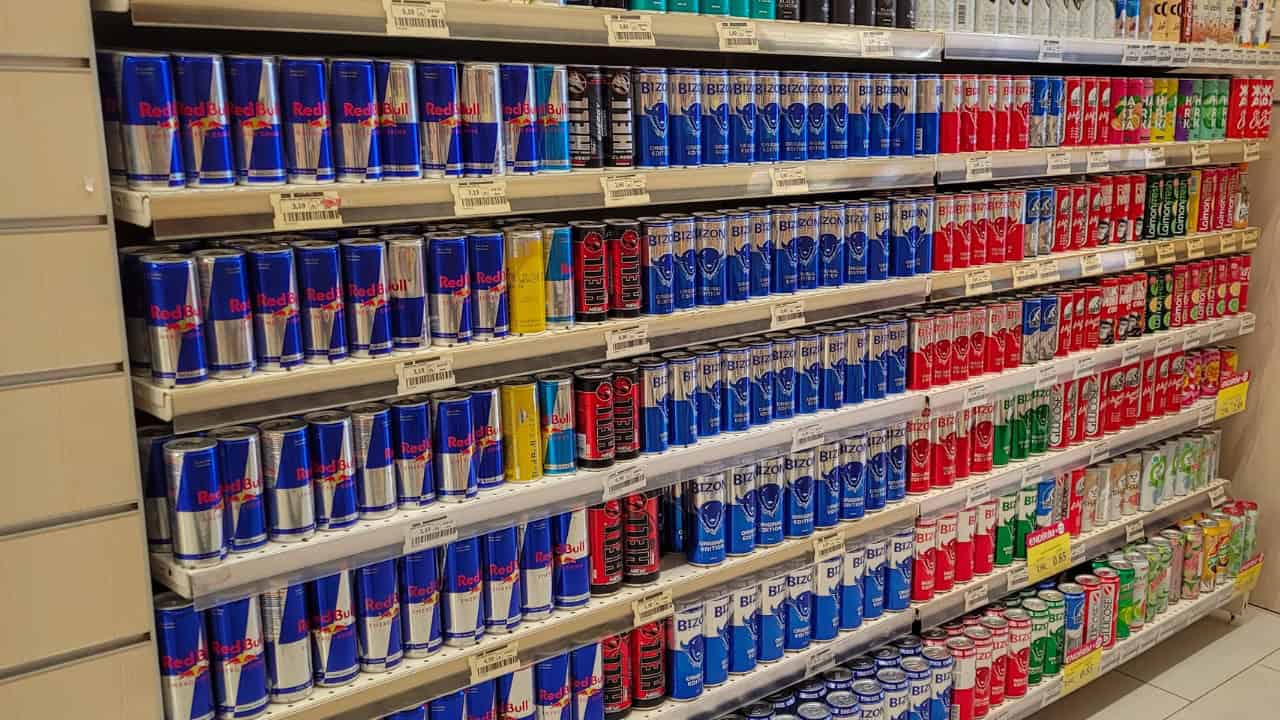 The image shows a shelf filled with rows of Red Bull energy drink cans in various colors, neatly organized by type, some cans are arranged by size, different flavors and variants are visible, the background is a typical store aisle with shelves stacked with more cans