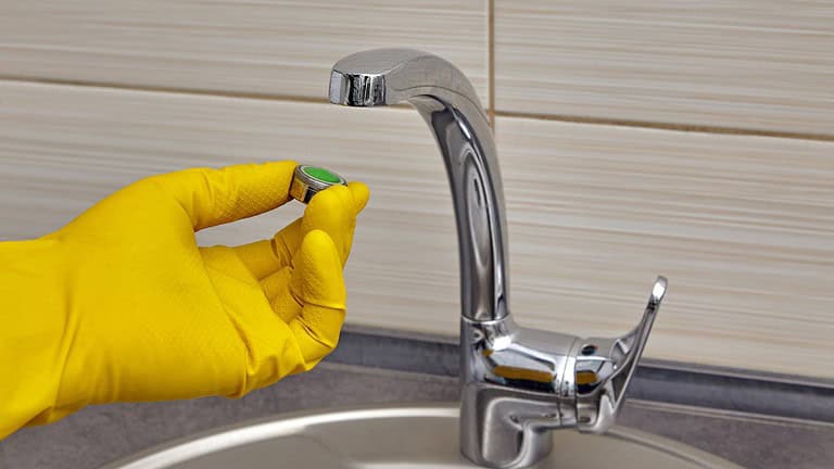 Person wearing yellow rubber glove cleaning a chrome kitchen faucet, beige tiled backsplash, stainless steel sink visible, household cleaning in progress