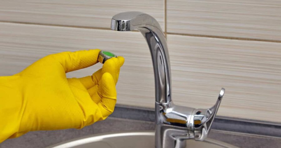 Person wearing yellow rubber glove cleaning a chrome kitchen faucet, beige tiled backsplash, stainless steel sink visible, household cleaning in progress