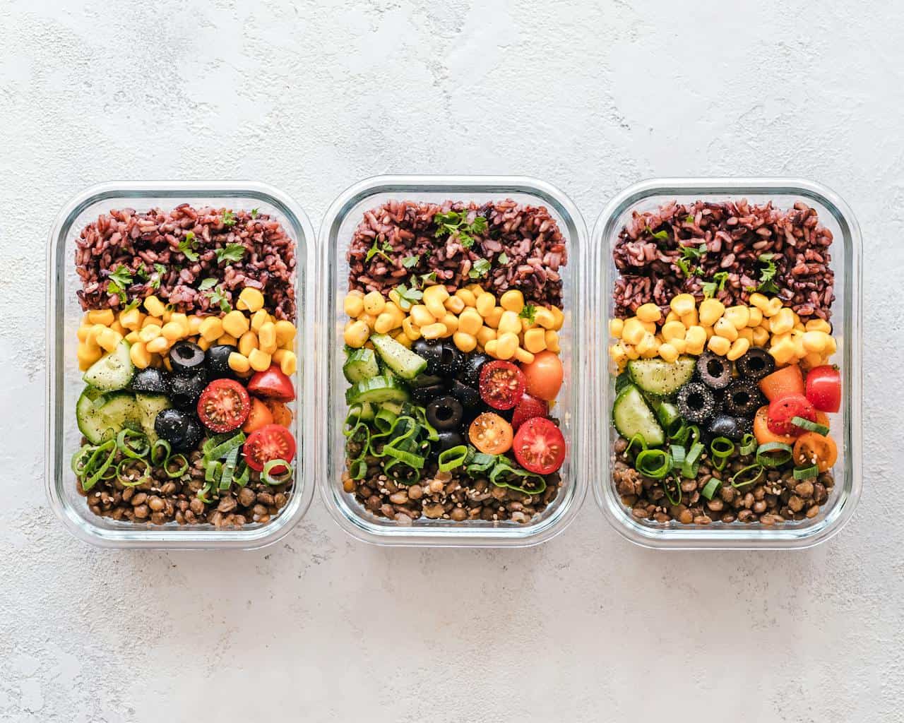 Three identical glass meal prep containers with layered vegetarian ingredients including purple rice, chickpeas, corn, black olives, cucumber, cherry tomatoes, and green onions, arranged on white surface