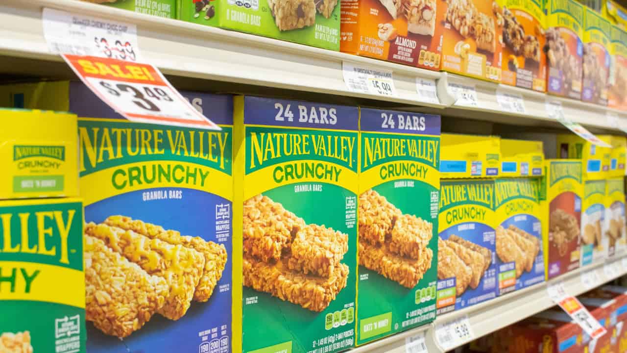 Boxes of Nature Valley Crunchy granola bars on a grocery store shelf, neatly arranged packaging, bright green and yellow boxes displaying product details, labels indicating 24 bars per box