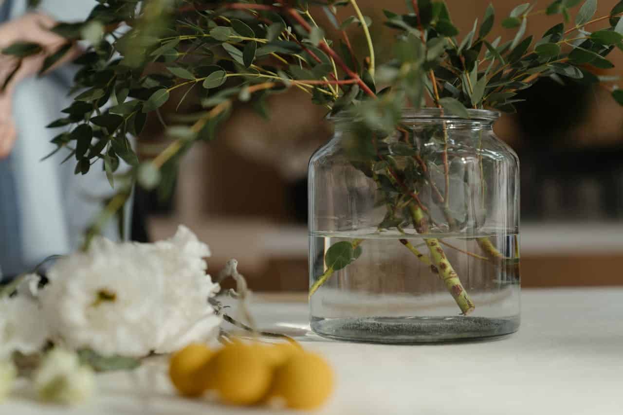 Glass jar with eucalyptus branches in water, white and yellow flowers nearby, soft-focus background, minimalist table arrangement
