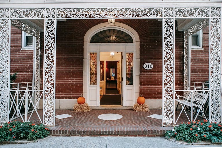 Brick house with ornate white ironwork, house number 310 displayed on the wall, double doors with glass panels, white benches on the porch, neatly arranged flowers, a welcoming entrance