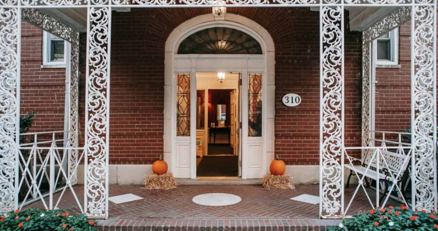 Brick house with ornate white ironwork, house number 310 displayed on the wall, double doors with glass panels, white benches on the porch, neatly arranged flowers, a welcoming entrance
