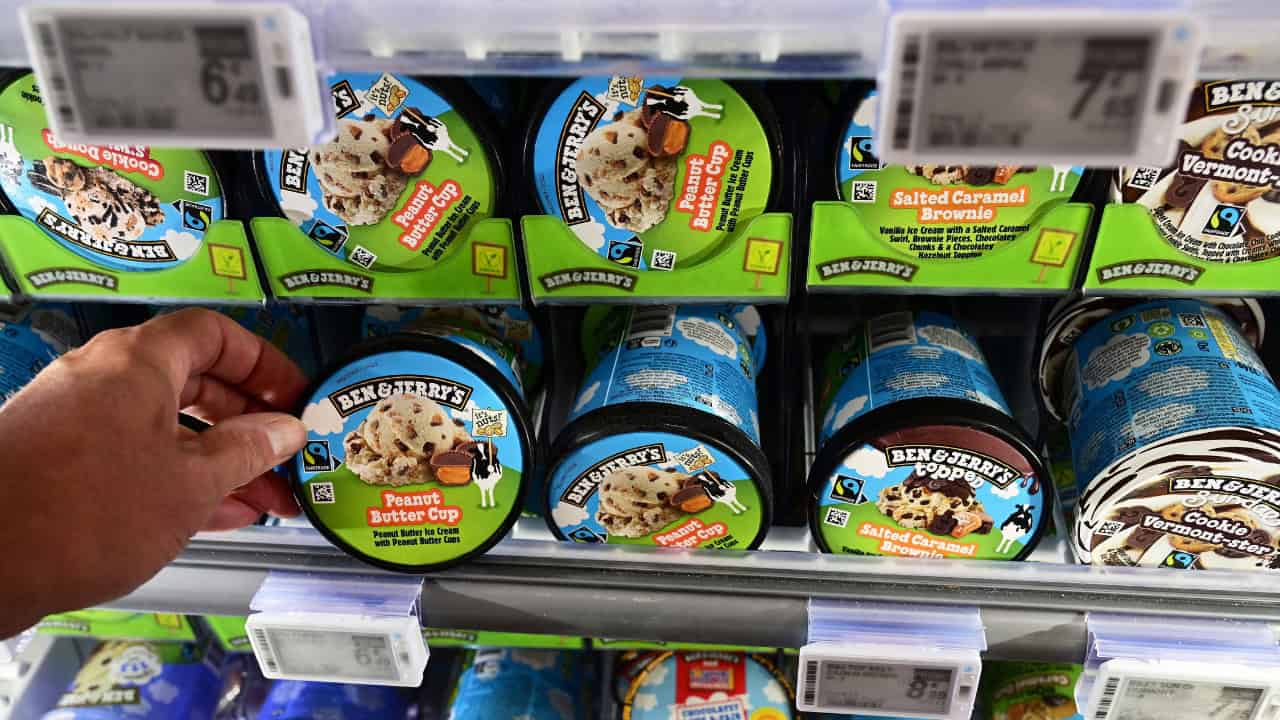 Ben & Jerry's ice cream tubs in a freezer, various flavors visible, a hand reaching for a tub, bright packaging with bold labels