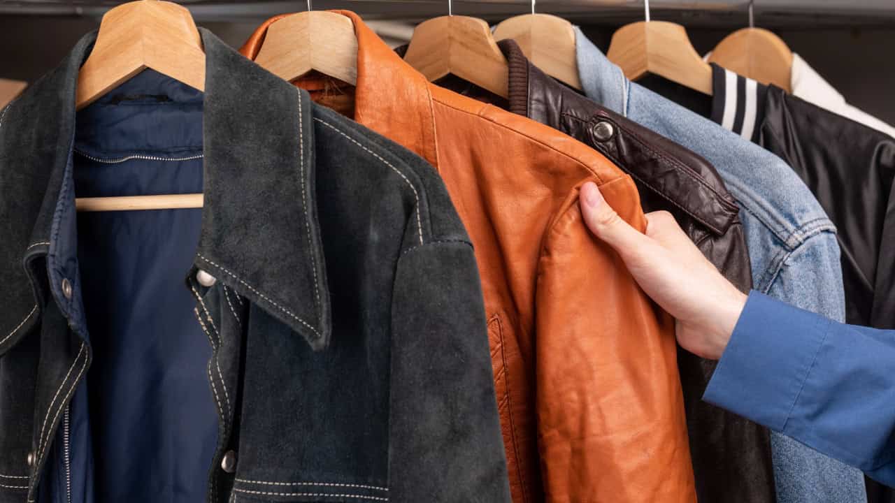 Clothing rack with leather jackets including a black suede jacket and an orange/brown leather jacket. Someone's hand is visible touching the orange jacket. A blue check icon appears in the corner