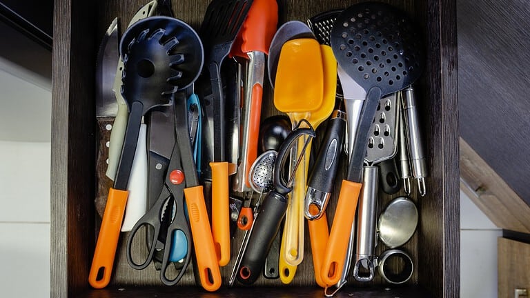 A collection of kitchen utensils, including spoons, spatulas, tongs, scissors, and measuring cups, with orange handles, placed in a disorganized manner in a drawer