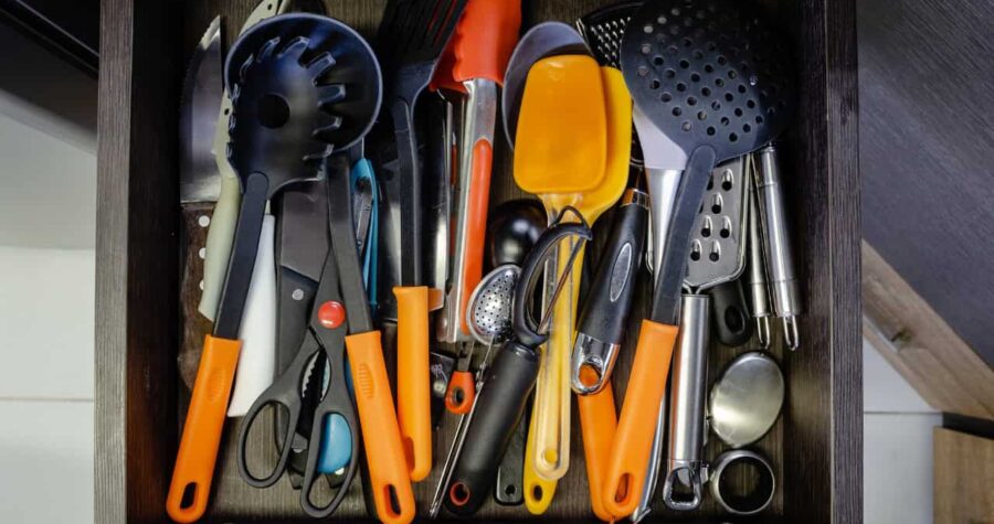A collection of kitchen utensils, including spoons, spatulas, tongs, scissors, and measuring cups, with orange handles, placed in a disorganized manner in a drawer