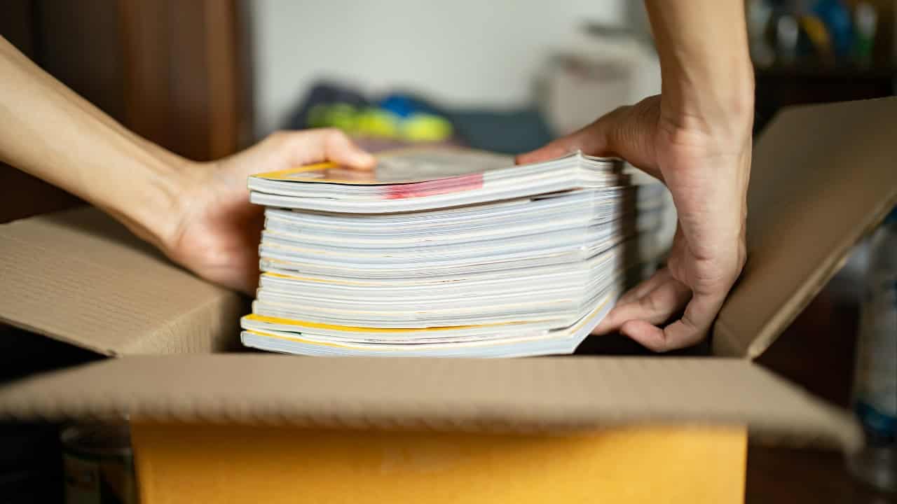 A person holding a stack of magazines, organizing magazines in a box, sorting through a pile of magazines, preparing magazines for storage, handling a stack of old magazines