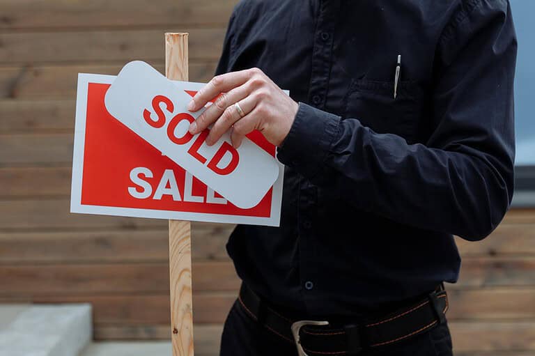 Person in black shirt placing a "SOLD" sticker over a "SALE" sign, wooden post, real estate signage, wooden background