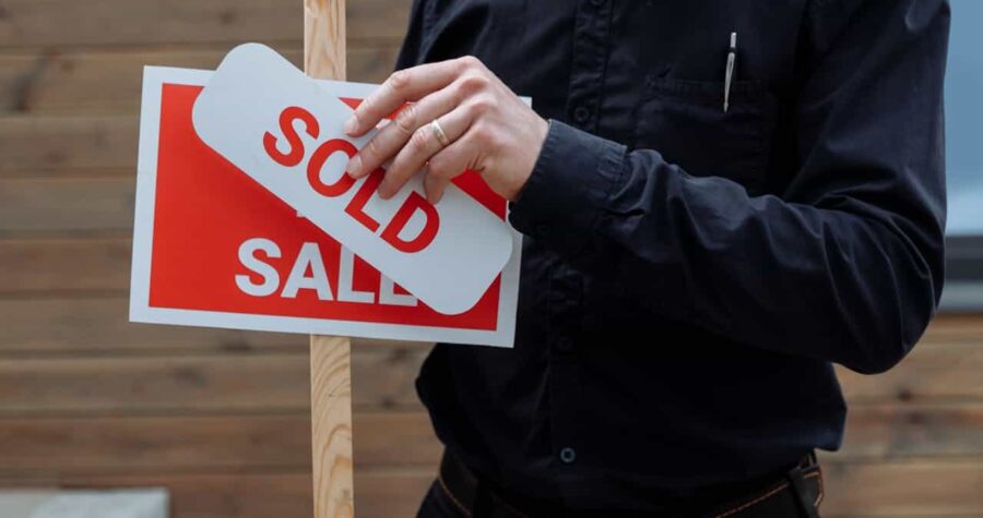 Person in black shirt placing a "SOLD" sticker over a "SALE" sign, wooden post, real estate signage, wooden background