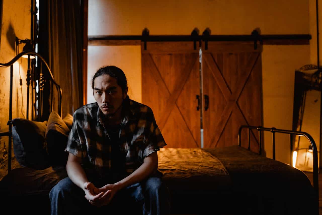 A person with long hair wearing a plaid shirt sits on the edge of a bed in a dimly lit room, warm orange lighting, sliding barn doors visible in the background, moody atmosphere, metal bed frame, rustic interior design