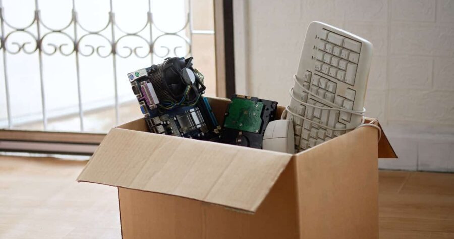 Cardboard box filled with old electronic components, including a keyboard, circuit boards, and computer parts, placed near a window, possibly for recycling or donation