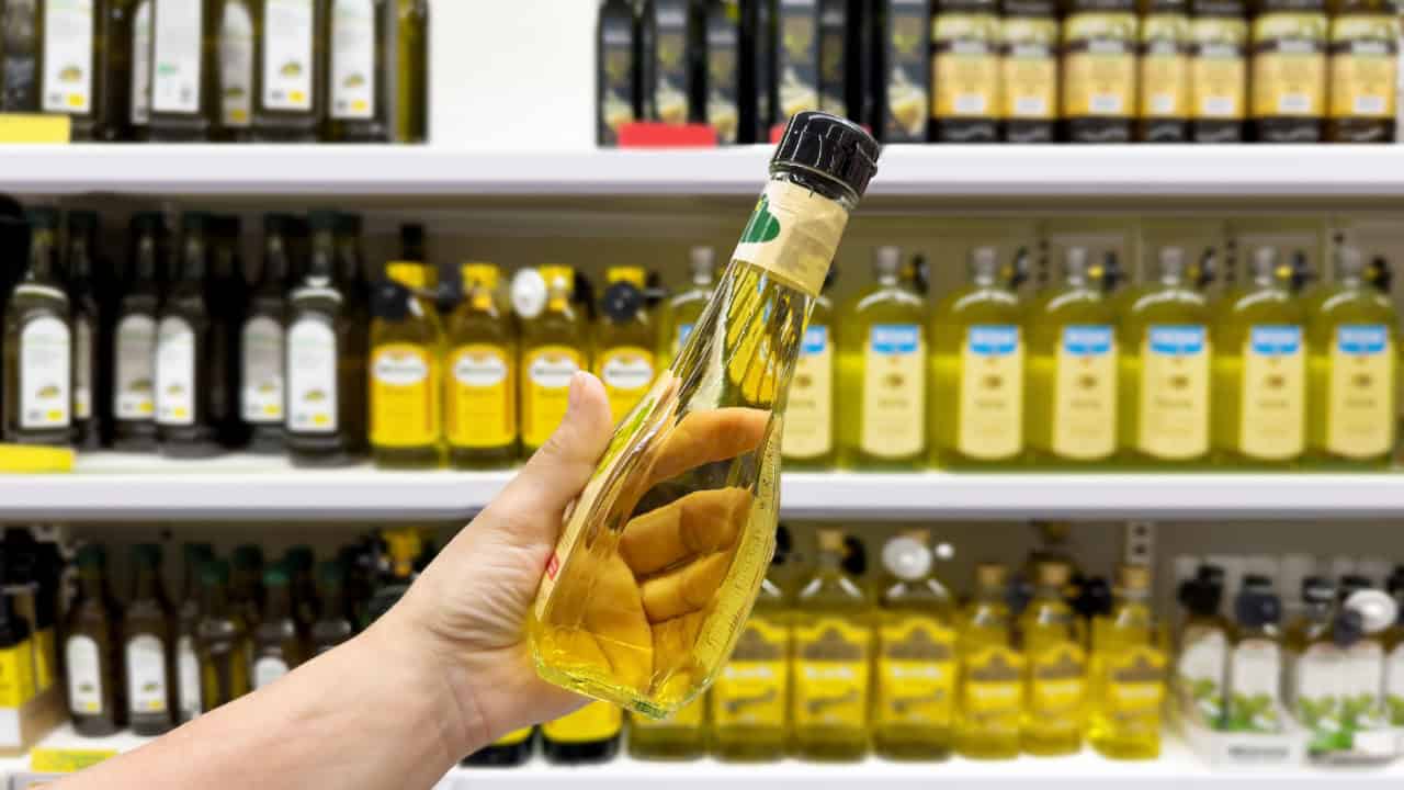 A person holding a bottle of olive oil, shelves of olive oil bottles in the background, grocery shopping for cooking oil, glass bottle with golden liquid