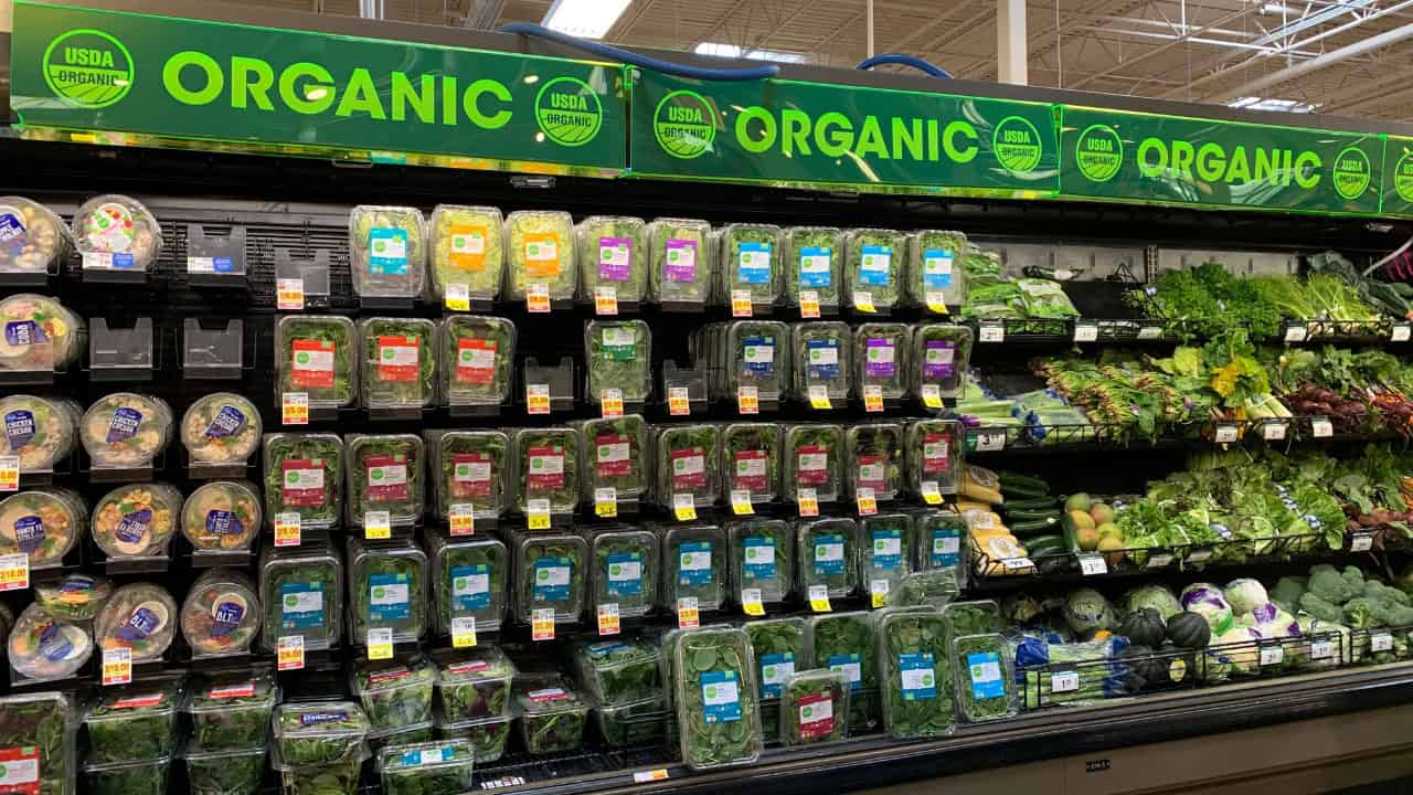 Organic produce section, neatly arranged leafy greens, various packaged salads, green, yellow, blue, and purple labels, brightly lit grocery store shelf, organized food display, emphasis on organic options