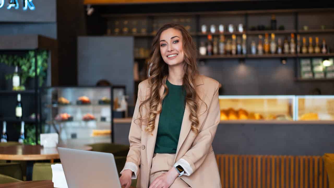 A smiling woman with long blonde hair wearing a beige or tan blazer sits in what appears to be a cafe or restaurant with warm lighting and shelving visible in the background