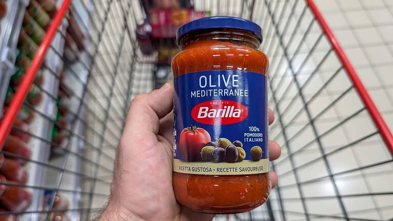 A jar of Barilla Olive Mediterranean pasta sauce held in a person's hand, displaying the label with a tomato, olives