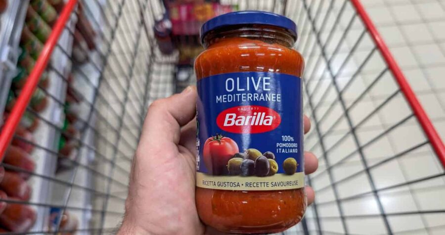 A jar of Barilla Olive Mediterranean pasta sauce held in a person's hand, displaying the label with a tomato, olives