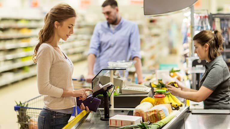 A woman scanning her items at a checkout counter, holding a payment card, grocery shopping, cashier station with produce and packaged goods on the counter