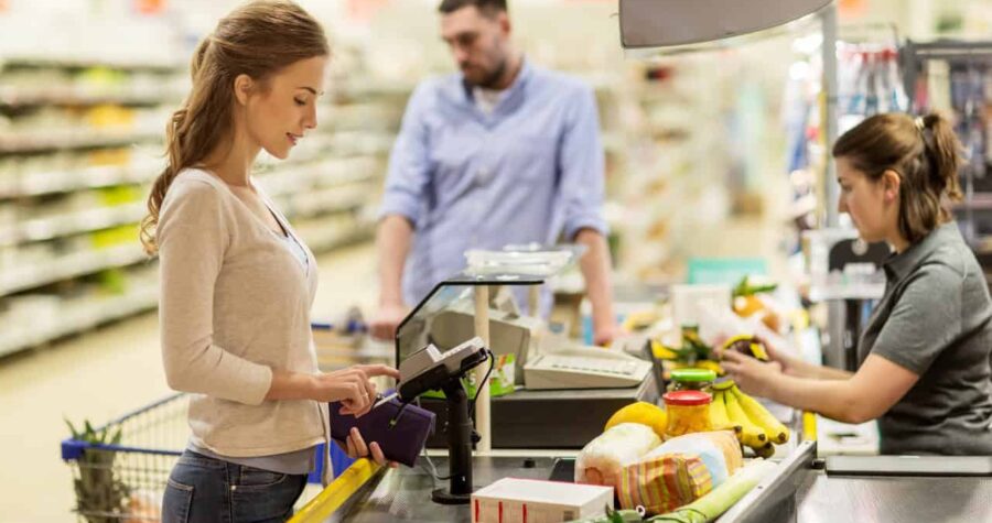 A woman scanning her items at a checkout counter, holding a payment card, grocery shopping, cashier station with produce and packaged goods on the counter