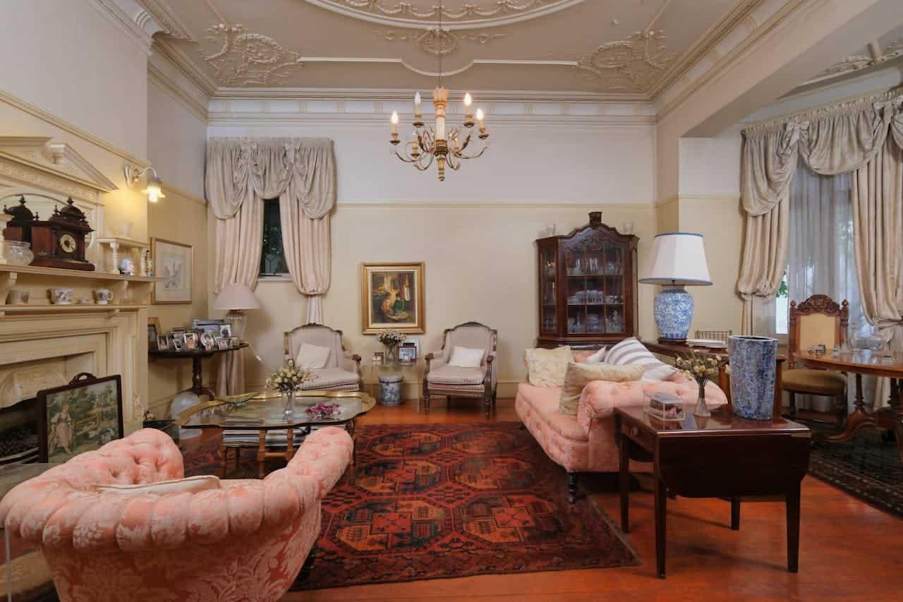 The image shows a classic, elegant living room with a vintage chandelier, plush pink seating, a patterned rug, and antique furniture, large windows with cream-colored curtains, framed artwork on the walls, a clock on the shelf, and decorative objects on the table