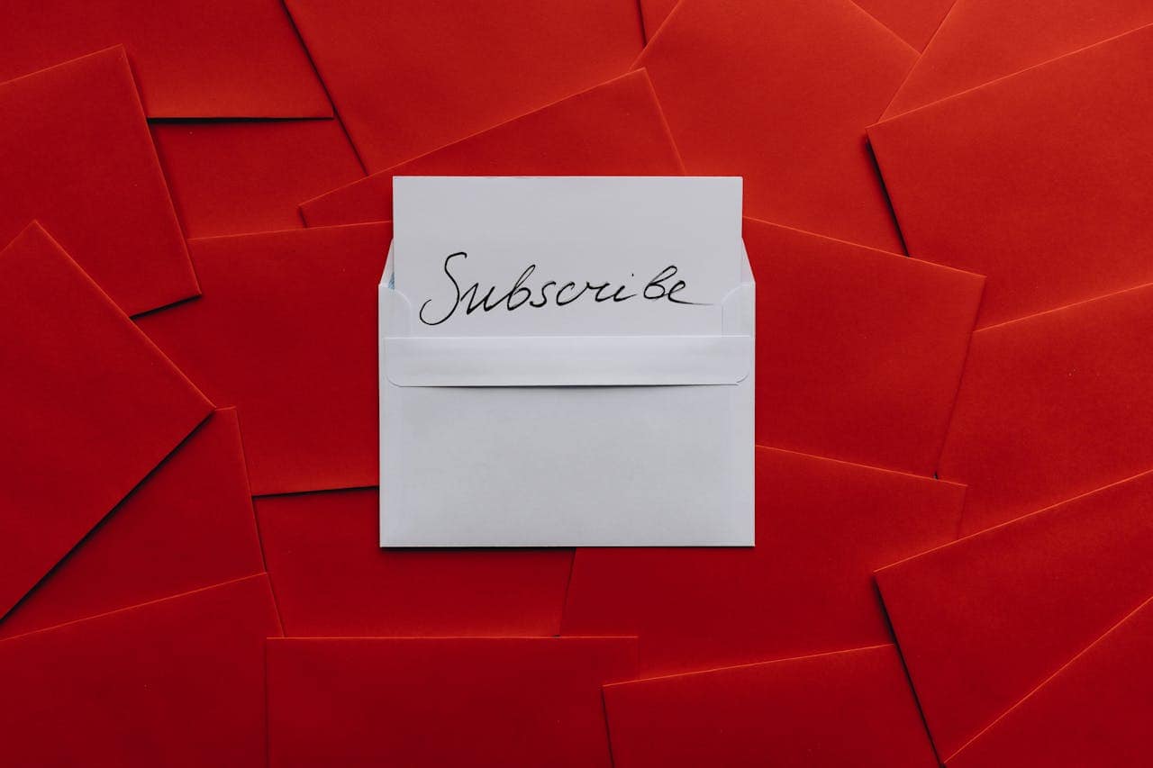 White envelope with "Subscribe" handwritten on it, surrounded by red envelopes, close-up shot, focus on the envelope and the word "Subscribe," vibrant colors, emphasis on a call to action or subscription theme, personal communication or marketing message