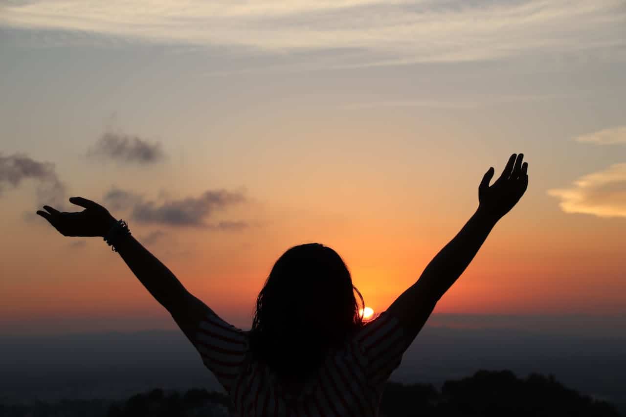 A person raising their arms in front of a sunset, silhouetted against a colorful sky, evoking a feeling of freedom, peace, and connection with nature, the warm colors of the sunset complementing the person’s joyful expression, standing outdoors, in a moment of tranquility