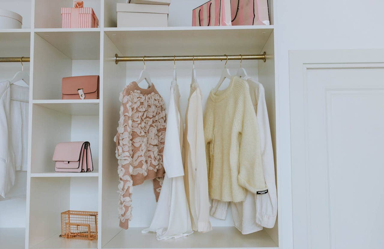 Organized white closet with pastel handbags, sweaters on hangers, pink gift boxes, and a small golden wire basket