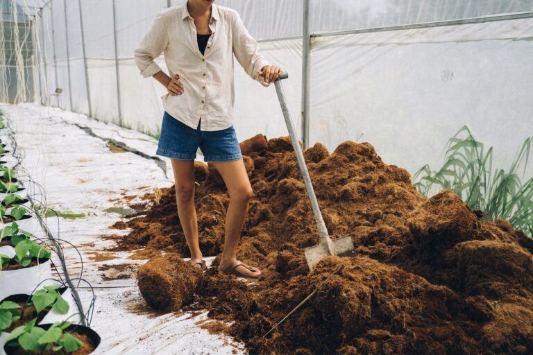 Person in white shirt and denim shorts working with shovel on brown mulch pile inside greenhouse with plants nearby