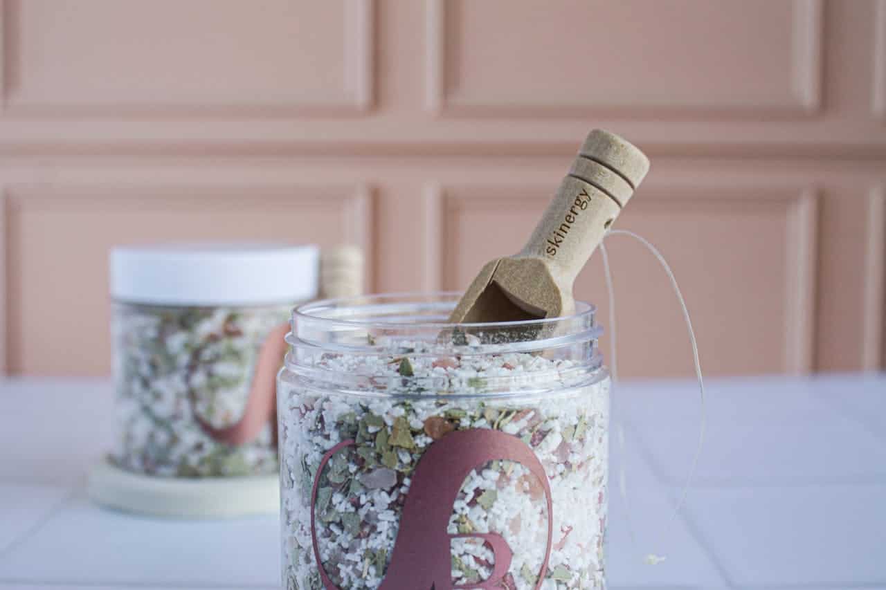 A jar of bath salts with a wooden scoop, the jar contains pink and white crystals mixed with dried herbs, the background features a soft peach-colored wall, another jar with similar contents is slightly blurred in the background