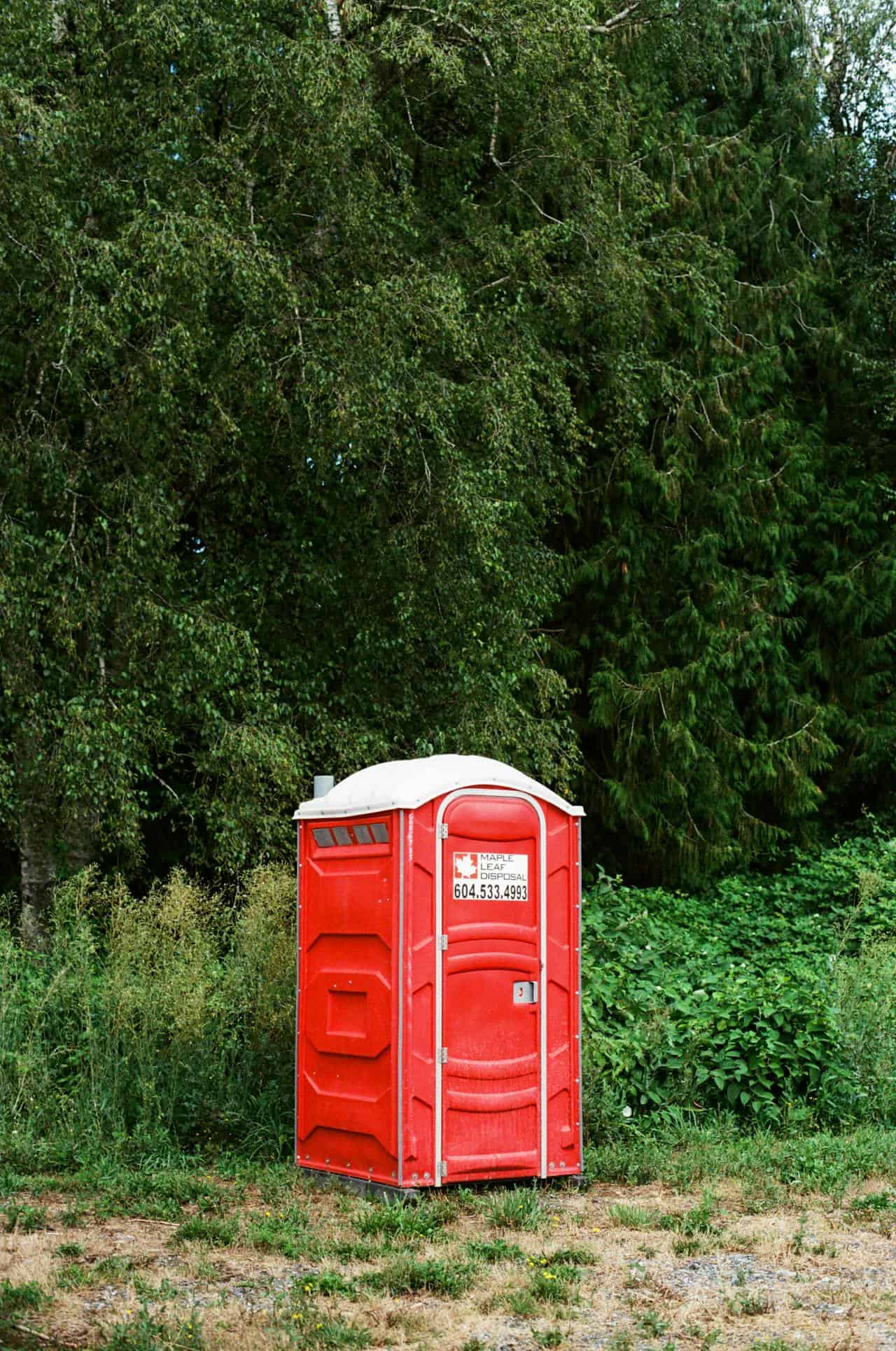 Red portable toilet with a white roof, placed on a patch of grass near dense green foliage and tall trees