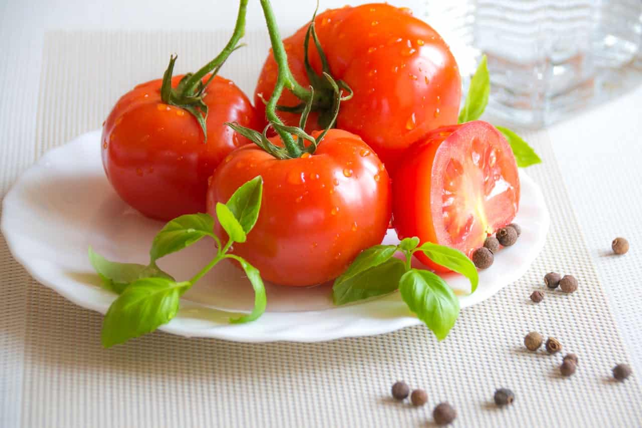 Fresh, ripe tomatoes, basil leaves, cut tomato on white plate, droplets of water on tomatoes, black peppercorns scattered, bright red tomatoes, healthy vegetables