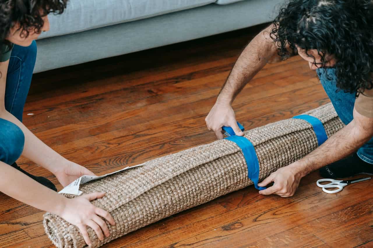 Two people are rolling up a carpet, securing it with blue tape, they are working together to roll the rug tightly, they are preparing the carpet for storage