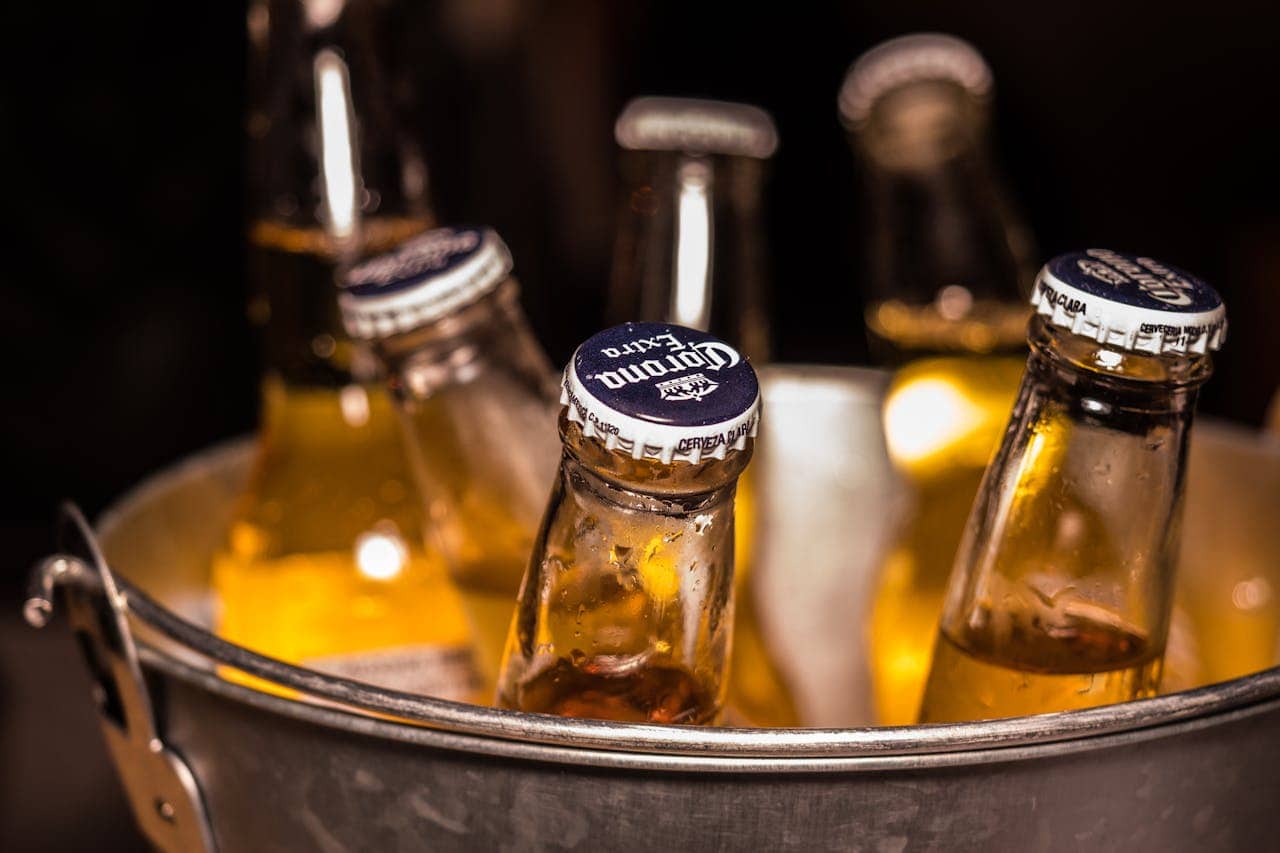 Beer bottles with Corona Extra labels in a metal bucket, bottles with caps visible, chilled beers in a bucket, close-up of beer bottles, refreshing alcoholic beverages, bottles of Corona with condensation