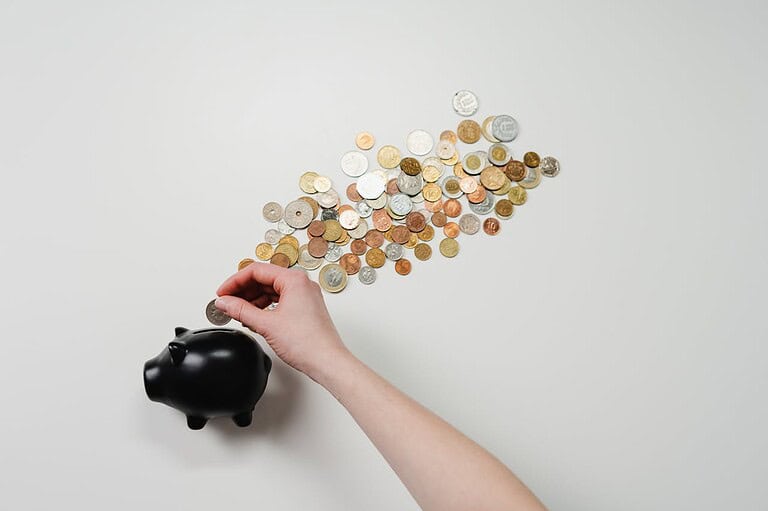 A hand placing a coin into a black piggy bank, scattered coins arranged in a trail around the piggy bank, the image illustrates saving money, coins of different denominations, financial management