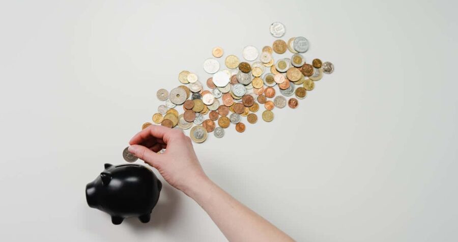 A hand placing a coin into a black piggy bank, scattered coins arranged in a trail around the piggy bank, the image illustrates saving money, coins of different denominations, financial management
