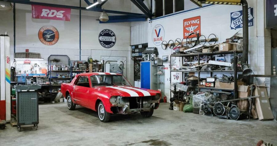 Vintage red Ford Mustang with white racing stripes being restored in auto shop with vintage signs and tools