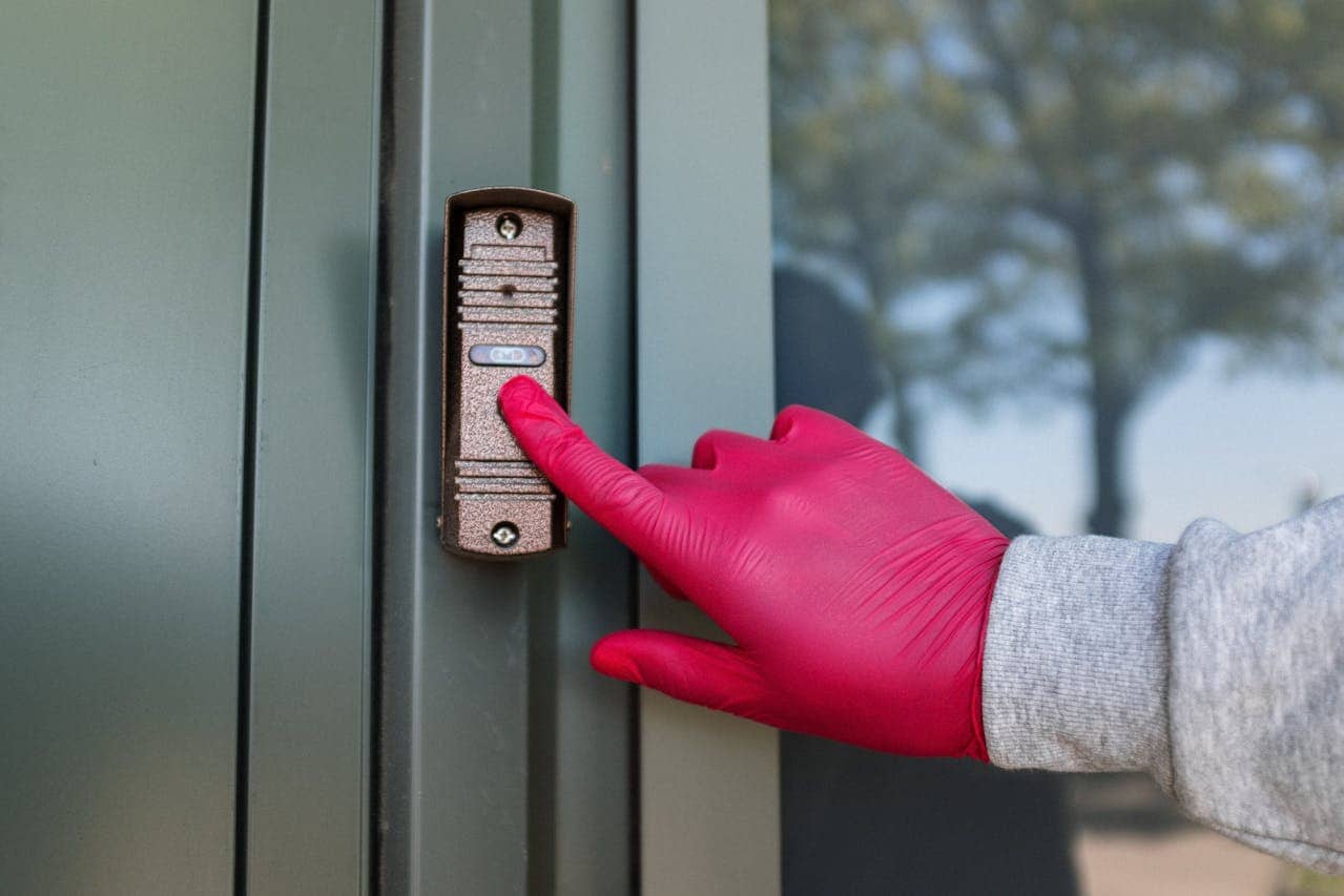 A person wearing pink gloves presses a doorbell on a green door, the action suggests an attempt to call or request entry, the background shows blurred greenery, the person is wearing a gray sweatshirt, the scene is clear and focuses on the hand pressing the button