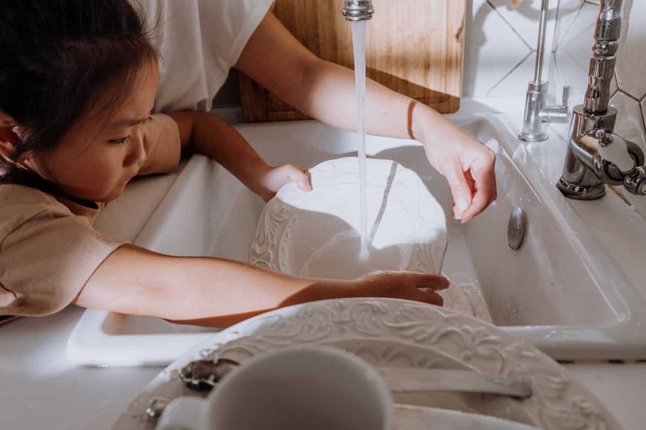 A child washing dishes with an adult, hands in a sink, washing a white plate, water flowing from the faucet, helping with chores