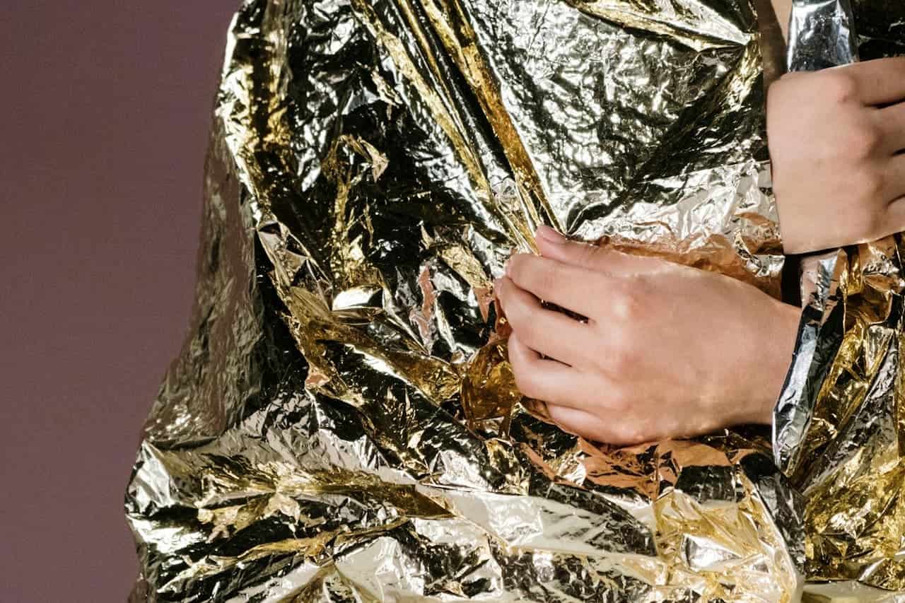 Person wearing Emergency Mylar Blankets wrapped around their body, with hands visible against a dark burgundy background