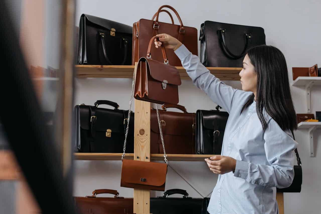 A person is holding a brown handbag, several other leather bags are visible on the shelves in the background, the person is inspecting the bag