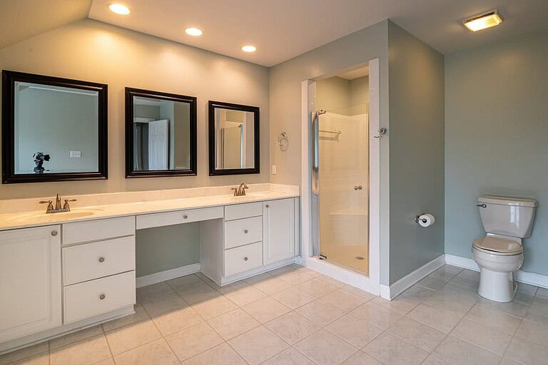 Spacious bathroom with a long white vanity, three black-framed mirrors, overhead recessed lighting, enclosed glass shower, tiled floor, and light gray walls